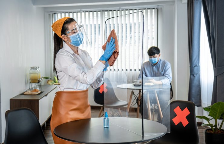 Asian woman waitress wearing protective face mask and medical gloves while using alcohol to wipe table plastic shield partition for cleaning before serving customers in cafe