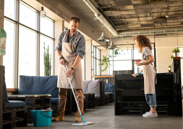 Busy young couple in aprons washing floor and cleaning surfaces with detergent while preparing cafe before opening